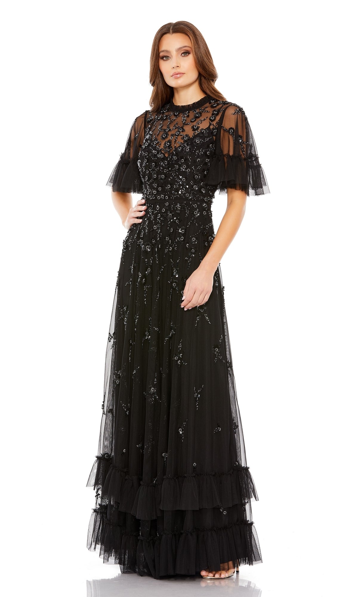 A woman stands against a white background in the Mac Duggal 9199 Short Sleeve Beaded Formal Gown, featuring sheer short sleeves, floral embellishments, and a layered tulle skirt.